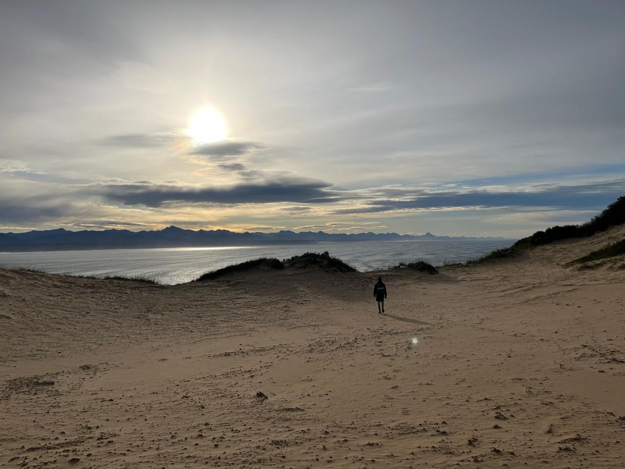 A lone figure walking across a coastal dune at sunset, with the ocean and mountain range beyond